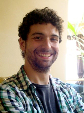 Man with dark curly hair wearing a plaid shirt and smiling into the camera
