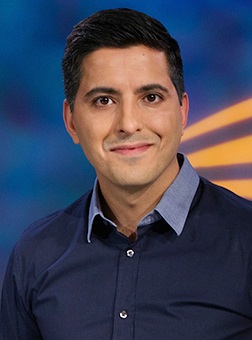 Image of a man with dark hair wearing a navy shirt. He is standing against a blue background and smiling into the camera