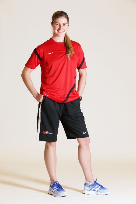 Woman wearing a red shirt and black shorts and trainers with her hands in her pockets and smiling into the camera